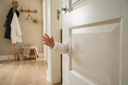 Enfant en bas âge approchant ses doigts du bord d'une porte en bois blanche dans un couloir familial, illustrant le risque de pince-doigt